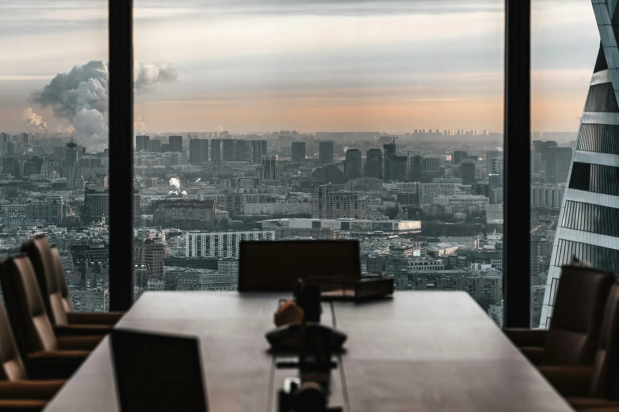 A darkened boardroom table facing floor-to-ceiling windows and a hazy city skyline at dusk