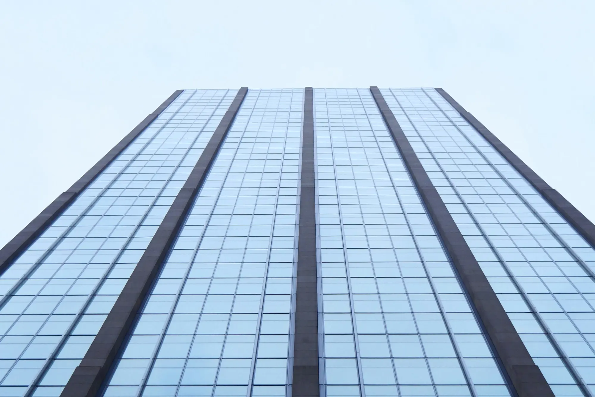 A glass curtain-wall office tower viewed from below against a pale sky
